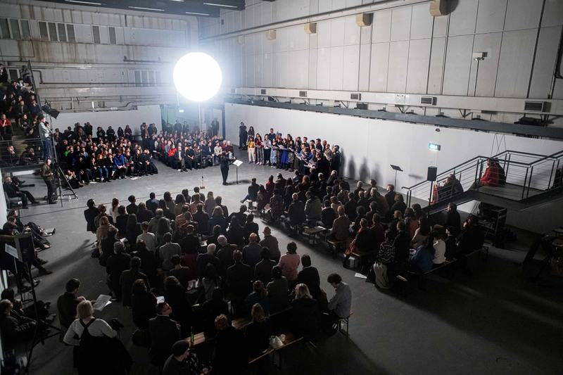 A large audience gathers around an enormous bright weather balloon in an underground space – a choir sings