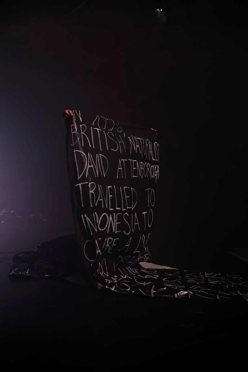 A man holding a black sheet of plastic with writing covering his entire body on stage