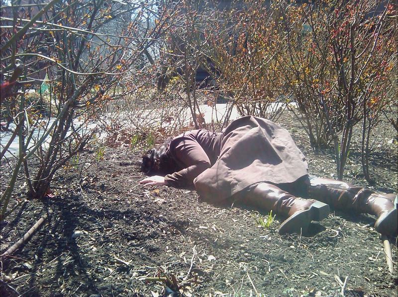 Poet Bhanu Kapil lies face down on the earth in a park in a coat and boots, surrounded by shrubs.